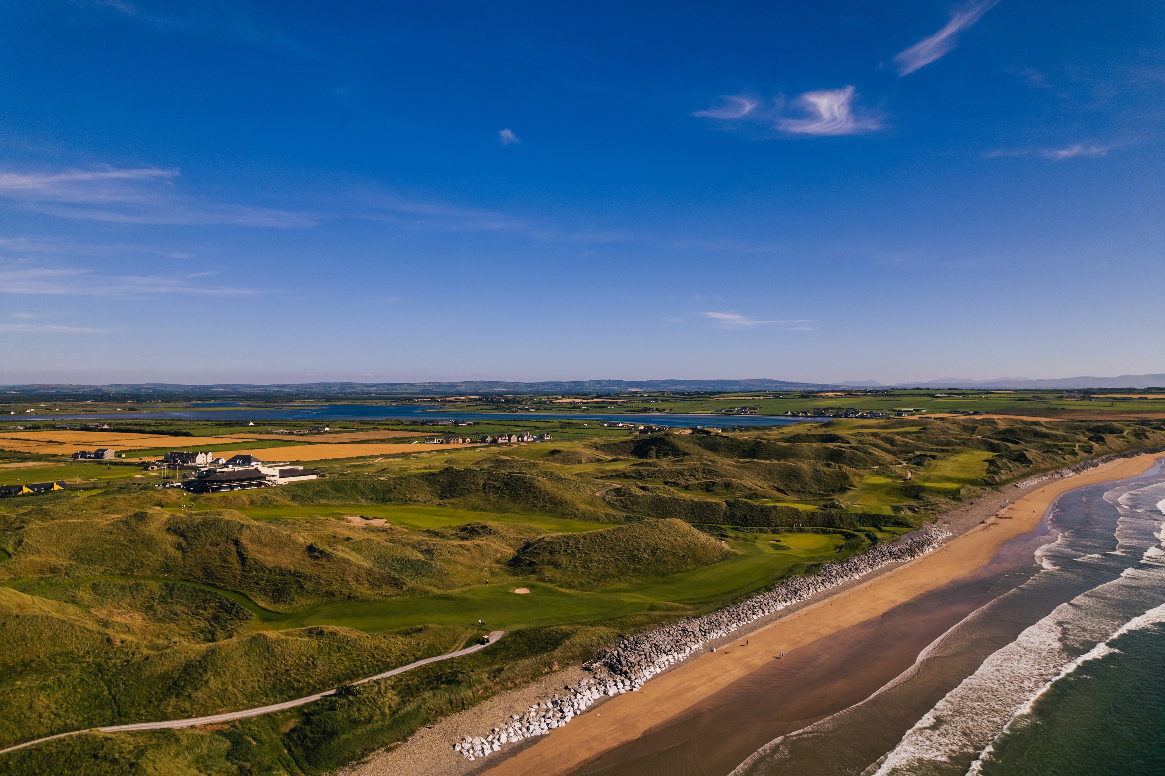 Aerial view of a coastal landscape with green fields, a beach, and a clear blue sky.
